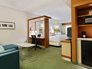 Hotel room with a bed, desk with chair, sofa, coffee table, TV, microwave, fridge, and coffee maker. Neutral colors and green carpet. Wall art and window with curtains in the background.