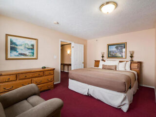 A bedroom with a large bed, wooden dresser, armchair, and two landscape paintings on beige walls.