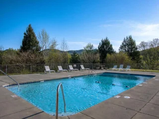 Outdoor swimming pool with clear water, surrounded by a concrete deck. Several lounge chairs are placed at the poolside. Trees and hills are seen in the background under a clear blue sky.