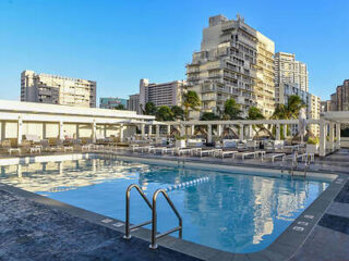 Outdoor swimming pool with lounge chairs and umbrellas, surrounded by tall buildings under a clear blue sky.