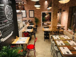 Cozy café interior with brick walls, wooden tables, and a mix of black, red, and gray chairs. Chalkboard menu on one wall and a plant in the corner.