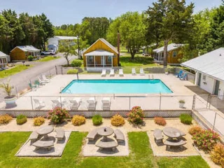 Aerial view of a community pool area surrounded by lounge chairs and small cottages, with landscaped greenery and picnic tables in the foreground.