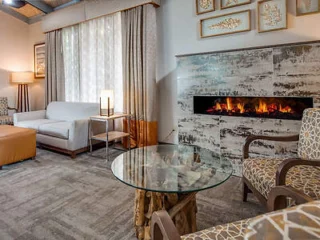 A cozy living room with a modern electric fireplace, a gray couch, patterned armchairs, and a round glass coffee table. Wall art and large curtains adorn the space.