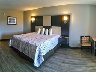 A hotel room with a king-sized bed, four pillows, a headboard with lights, a framed picture on the wall, and a chair with a desk on the side.