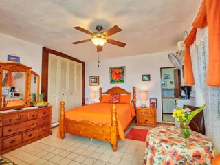 A cozy bedroom with a wooden bed, orange bedding, a ceiling fan, and floral decorations. A dresser with a mirror and a small table with flowers are also present.