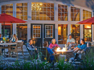 A group of people sit around a fire pit on a patio in front of a house with large windows.