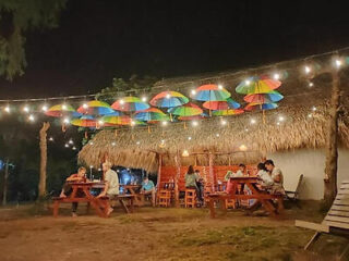 Outdoor cafe at night with people sitting at wooden tables. Colorful umbrellas hang above, and string lights illuminate the area.