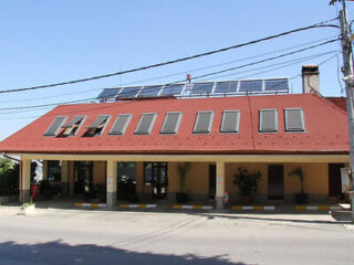 A building with a red sloped roof featuring solar panels and skylights, situated along a street with overhead power lines.