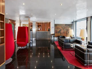 Modern lobby with black seating, red accents, and large windows. Unique high-back red chairs and a small bar area in the background.