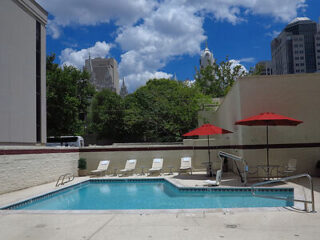 Outdoor pool area with lounge chairs and three red umbrellas, surrounded by buildings and greenery under a partly cloudy sky.