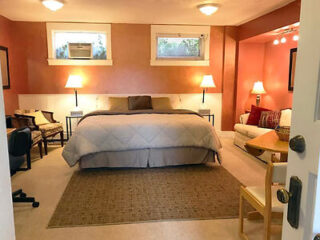 Cozy bedroom with a large bed, two side lamps, a sofa, and chairs. The walls are painted warm orange, and there are two small windows above the bed.