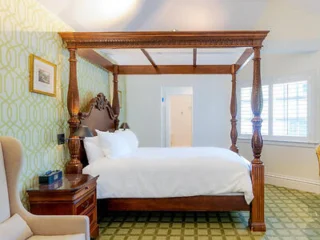 A room with a four-poster bed, wooden nightstand, armchair, patterned green wallpaper, and a window with blinds.