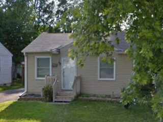 Small beige house with a gray roof, two windows, and a front porch with steps. Surrounded by trees and grass.