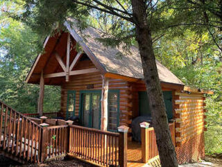 A small wooden cabin with a sloped roof, surrounded by trees. There is a wooden deck with a railing in front of the cabin.