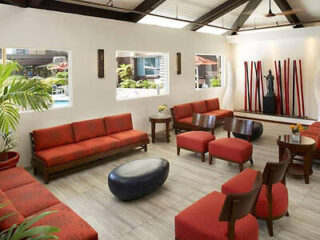 A brightly lit lounge with red sofas and chairs, a dark oval coffee table, and decorative wooden rods on the wall. Three windows provide natural light.