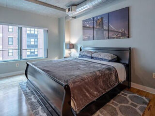 A modern bedroom with a large bed, gray bedding, and a cityscape photo on the wall. Large window with a view of buildings, hardwood floor, and area rug.