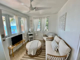 A small, bright living room with white walls, wicker furniture, a TV, and a ceiling fan. Ocean view through windows.