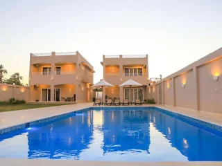 Two modern beige buildings with balconies surround a rectangular pool. Lounge chairs and umbrellas are placed on the poolside patio. The scene is set at dusk.