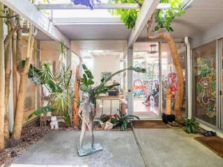 Indoor courtyard with a skylight, featuring a small statue, potted plants, and trees. Glass doors lead to an interior room decorated with colorful artwork.