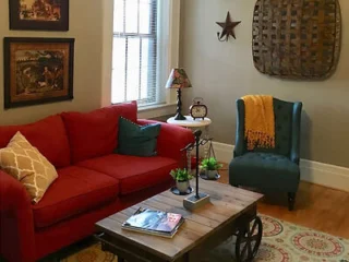 Cozy living room with a red sofa, blue armchair, wooden coffee table, and wall décor. A window with blinds allows natural light in. A small table with a lamp and clock sits by the sofa.