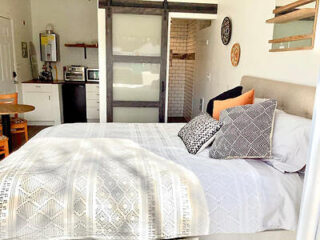 A cozy bedroom with a neatly made bed, several decorative pillows, a sliding door, and a small kitchenette in the background.