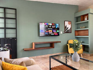 Living room with a wall-mounted TV, green painted walls, a wooden shelf, yellow armchair, glass coffee table, and decorative items on shelves.