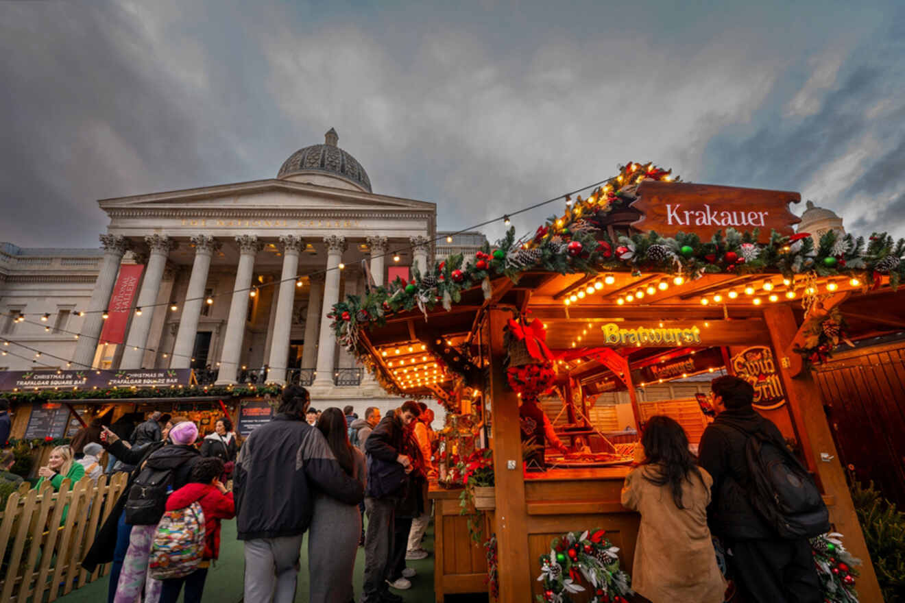 People gather at a festive market stall selling bratwurst in front of a grand building with columns under a cloudy sky.