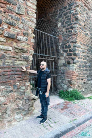 A person in sunglasses and casual attire stands with one hand on a brick and stone archway structure.