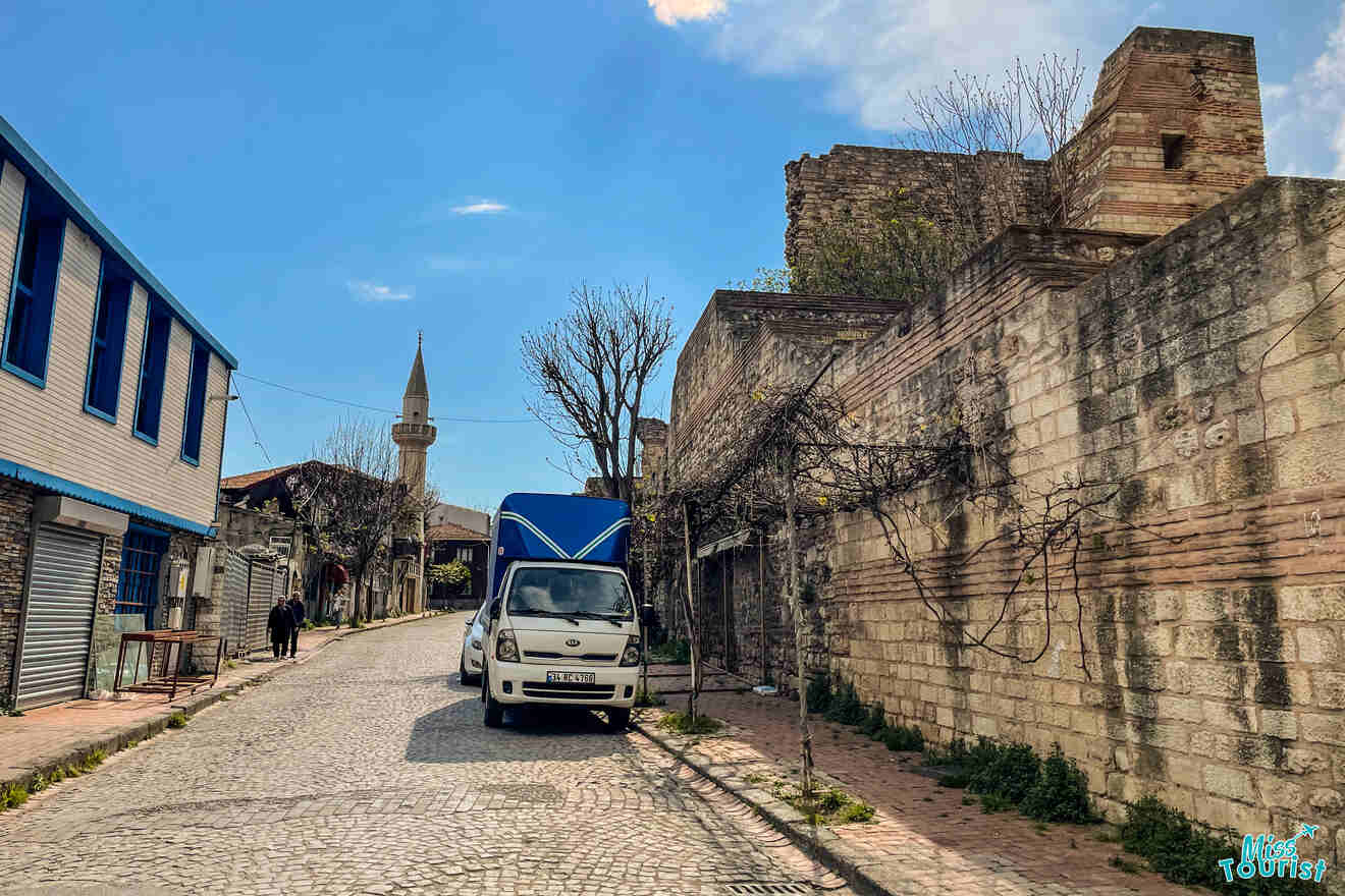 A narrow cobblestone street with parked van, lined by old stone buildings and bare trees. A minaret is visible in the background. Sparse pedestrian activity.