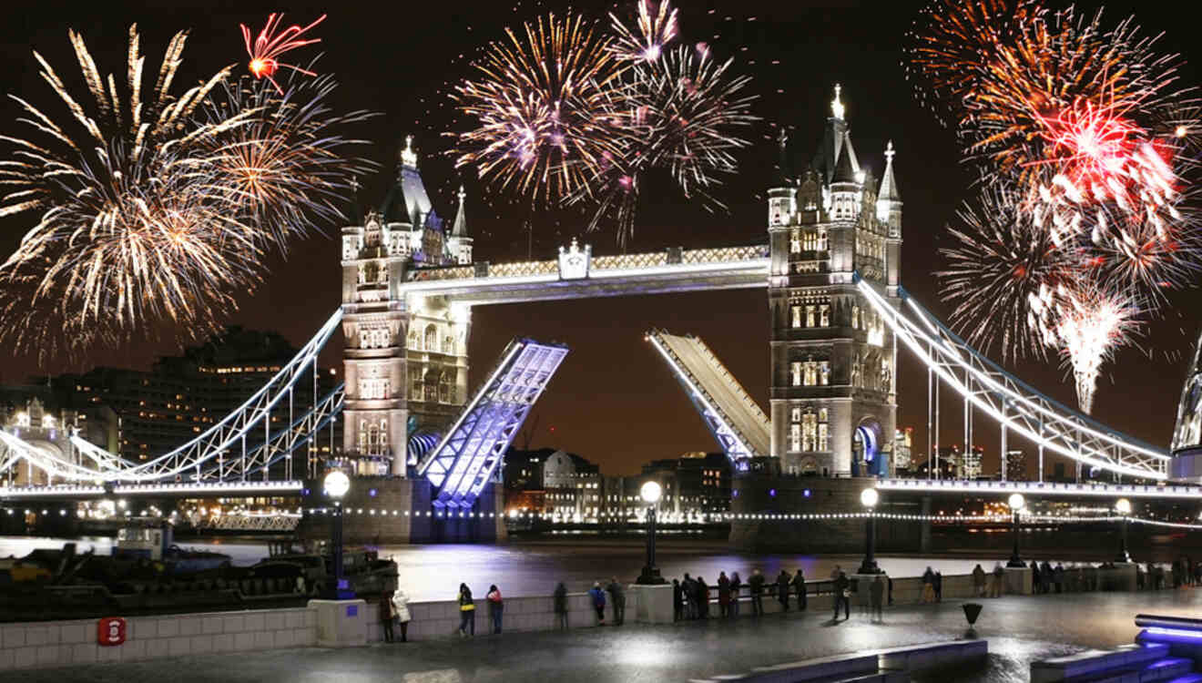 Fireworks illuminate the night sky above Tower Bridge in London, with the bridge raised and people watching from the riverside.