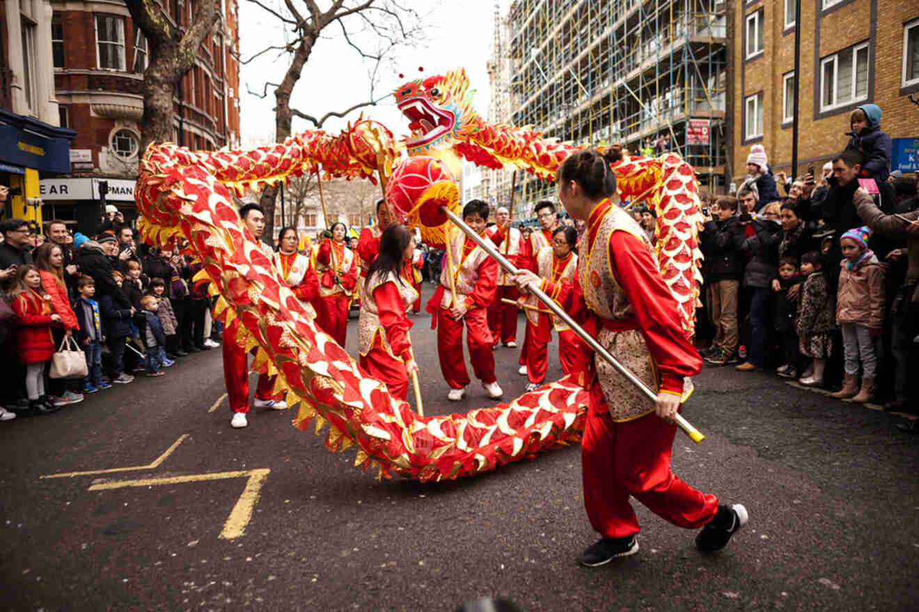 Performers in red and gold costumes participate in a dragon dance on a city street, surrounded by a crowd of onlookers.