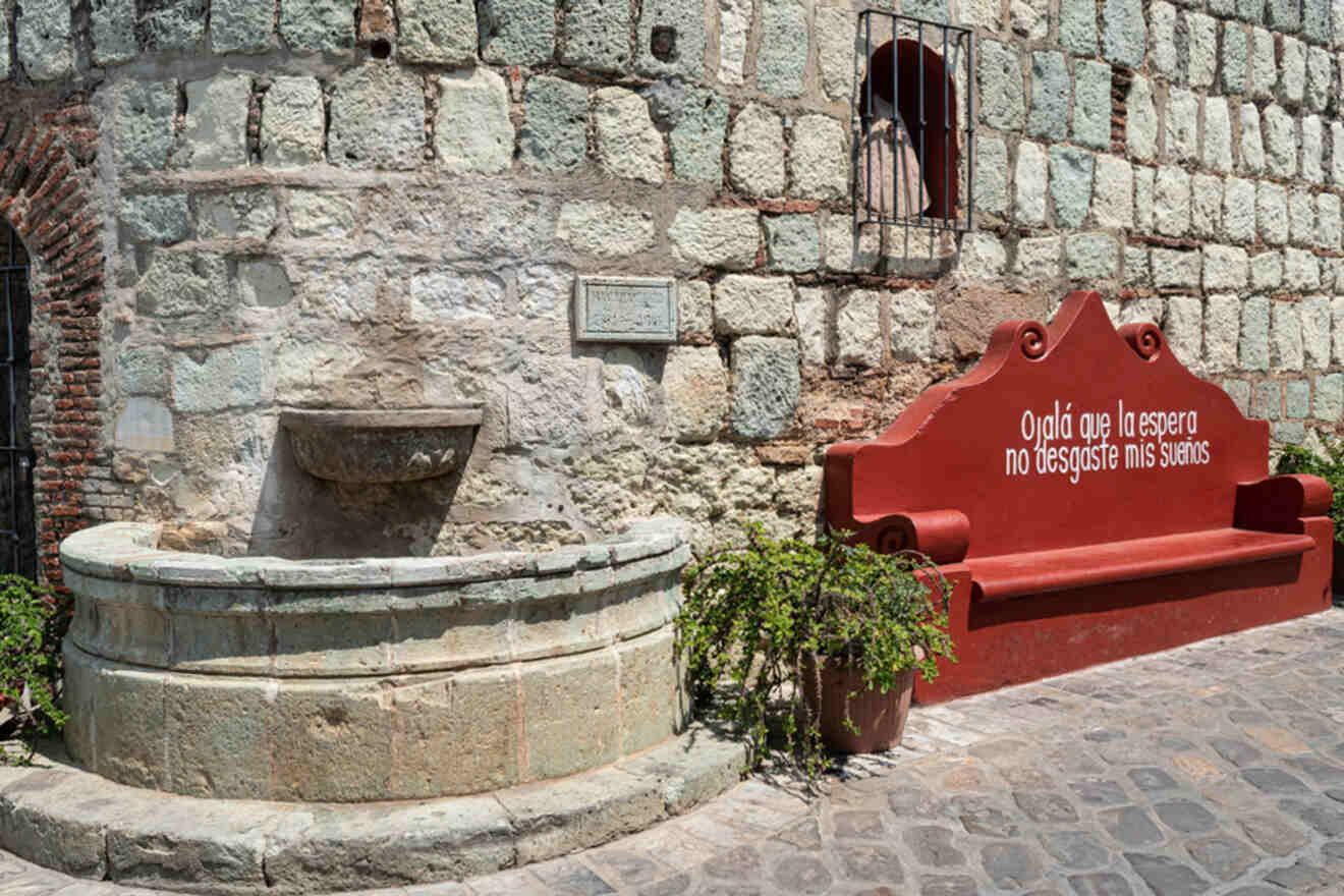 A stone wall with a round stone fountain beside a red bench. Spanish text on the bench reads: "Ojalá que la espera no desgaste mis sueños." Plants are placed near the fountain.