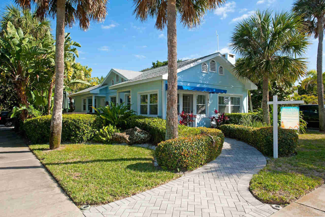 Charming light blue house with a gabled roof, surrounded by palm trees and manicured hedges, with a paved walkway leading to the entrance.