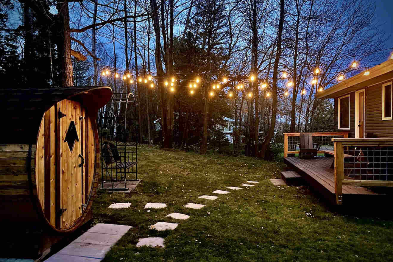 Backyard scene with a lit pathway, string lights above, a wooden sauna on the left, and a deck on the right at dusk.