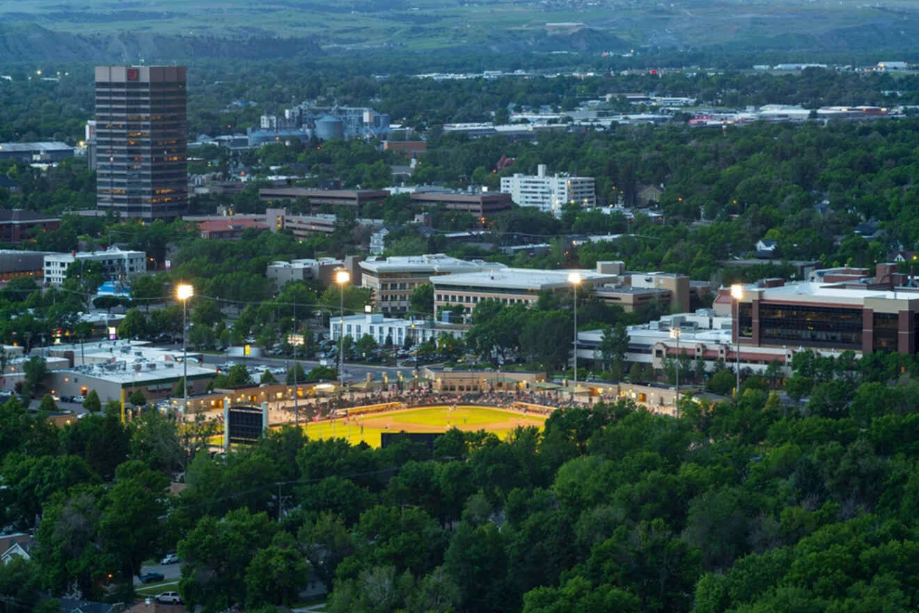 Aerial view of a baseball park lit up at dusk, surrounded by a cityscape with buildings, trees, and hills in the background.