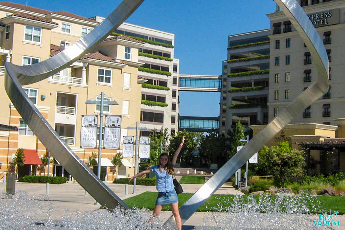 The writer of the post posing in front of a large metal sculpture surrounded by buildings and greenery.