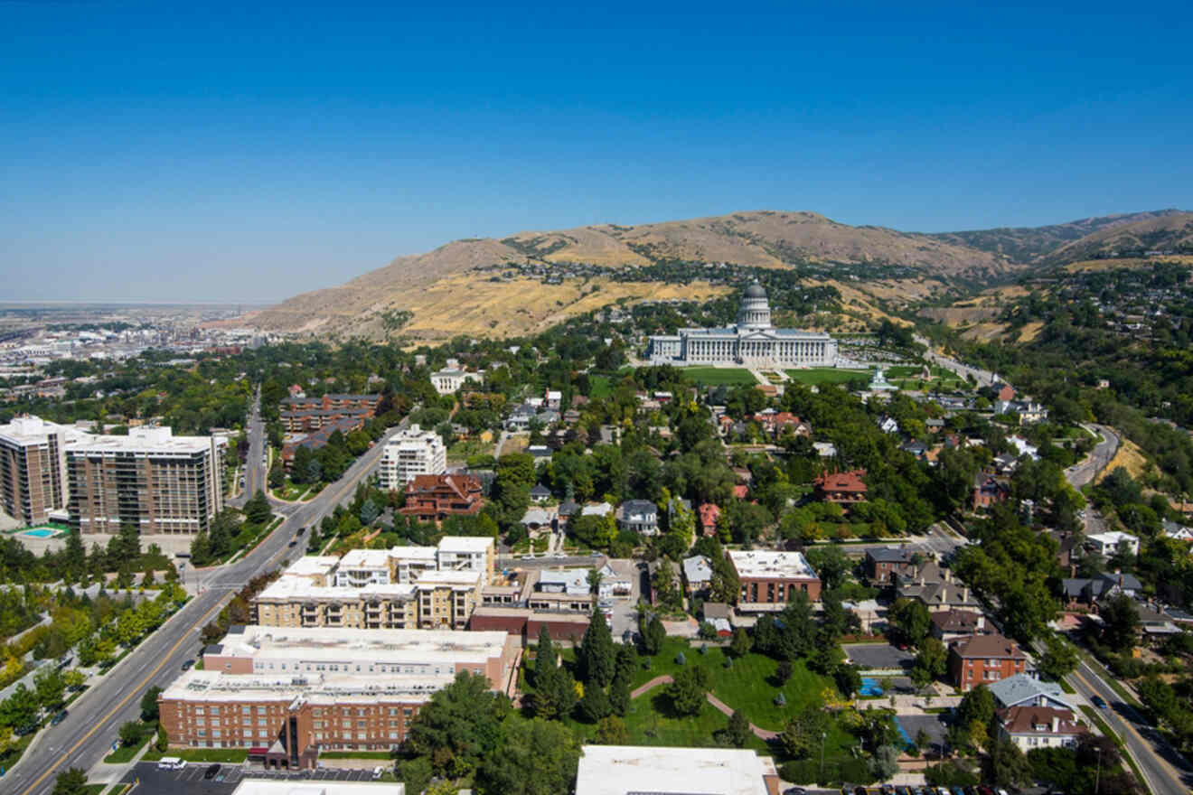 Aerial view of a city with a prominent white dome building, surrounded by greenery and hills under a clear blue sky.