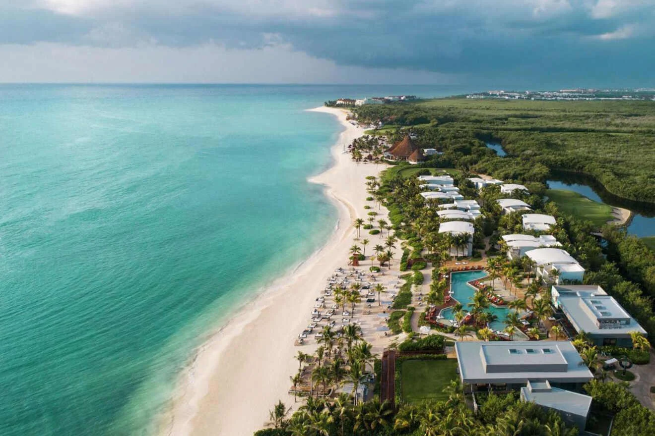 Aerial view of a tropical beach with turquoise water, a row of white beach chairs, palm trees, and a resort with pools next to a lush green landscape under a cloudy sky.