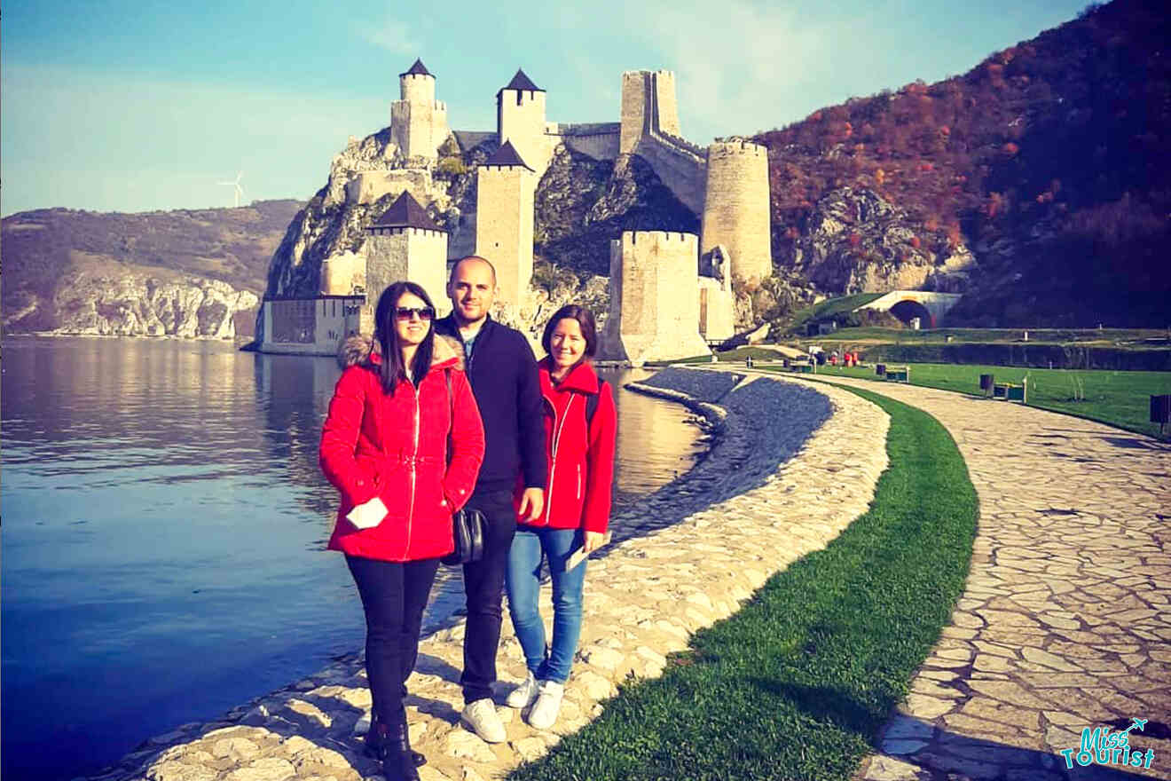 author of the post with her friends in red coats stand on a stone path by a tranquil body of water, with a medieval castle providing a stunning backdrop—an iconic stop on any Serbia itinerary.