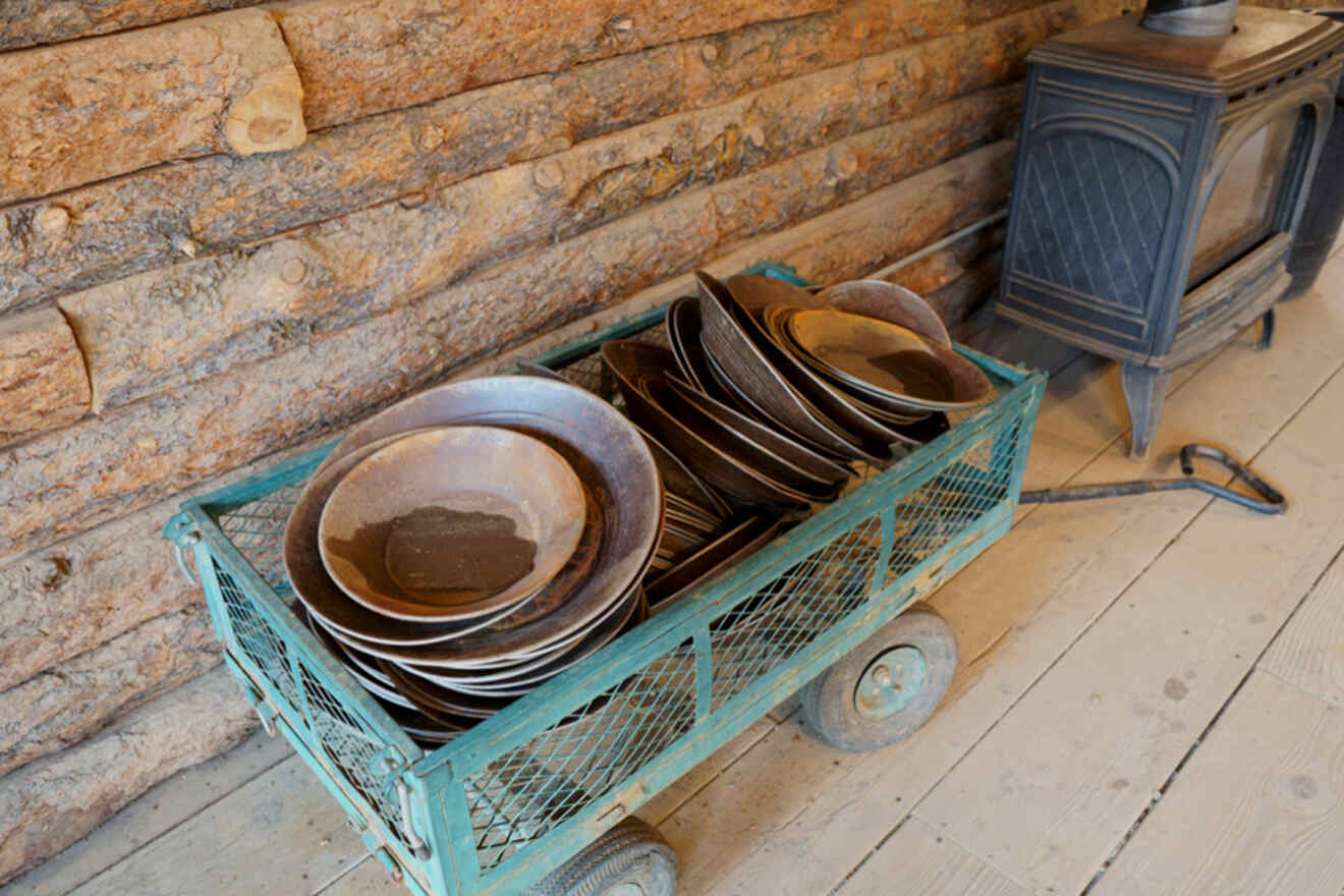 A teal metal wagon loaded with stacked brown ceramic plates is placed on a wooden floor next to a wood stove in a rustic setting.