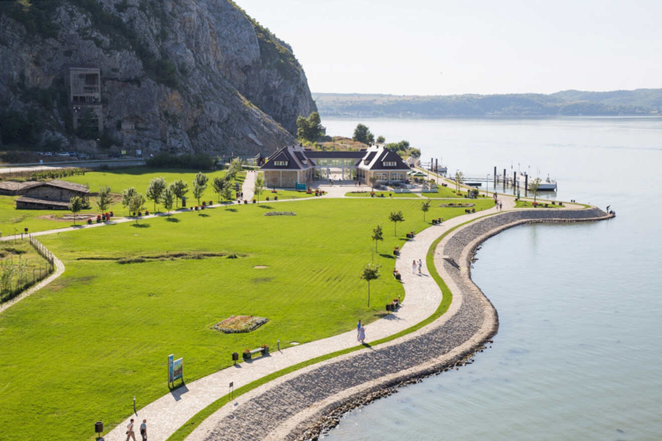 A scenic park with green lawns and walking paths curves along a waterfront. People stroll along the paths, with a hill and buildings in the background. A dock extends into the water.