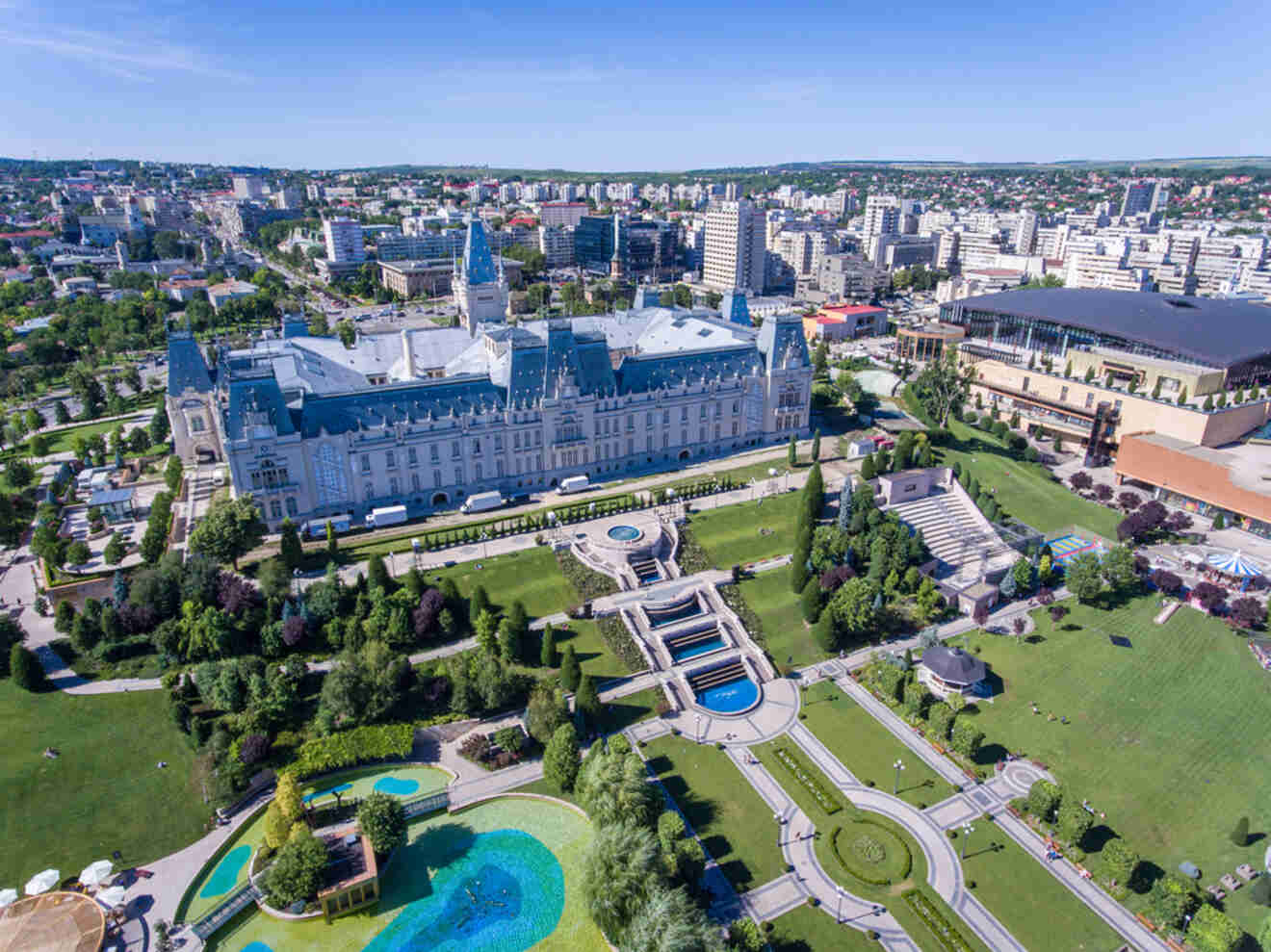 Aerial view of a large historical building with a clock tower, surrounded by landscaped gardens and fountains. The cityscape extends into the distance under a clear blue sky.