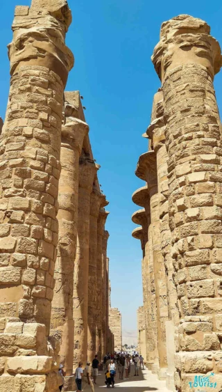 Tall ancient stone columns line a path under a clear blue sky, with tourists walking between them.