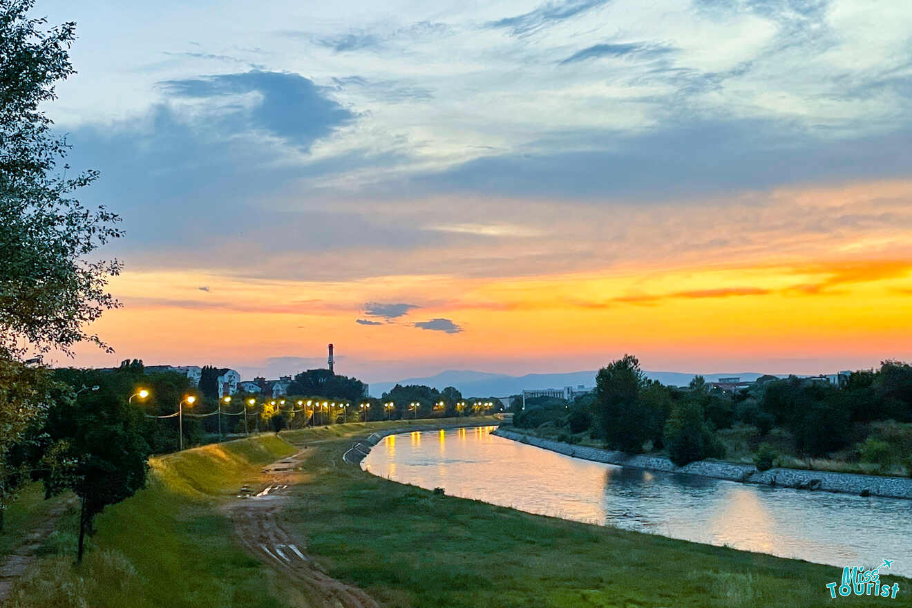 Sunset over a river, streetlights casting a warm glow along the path, trees swaying gently, and an industrial building silhouetted in the distance—a hidden gem for any Serbia itinerary.