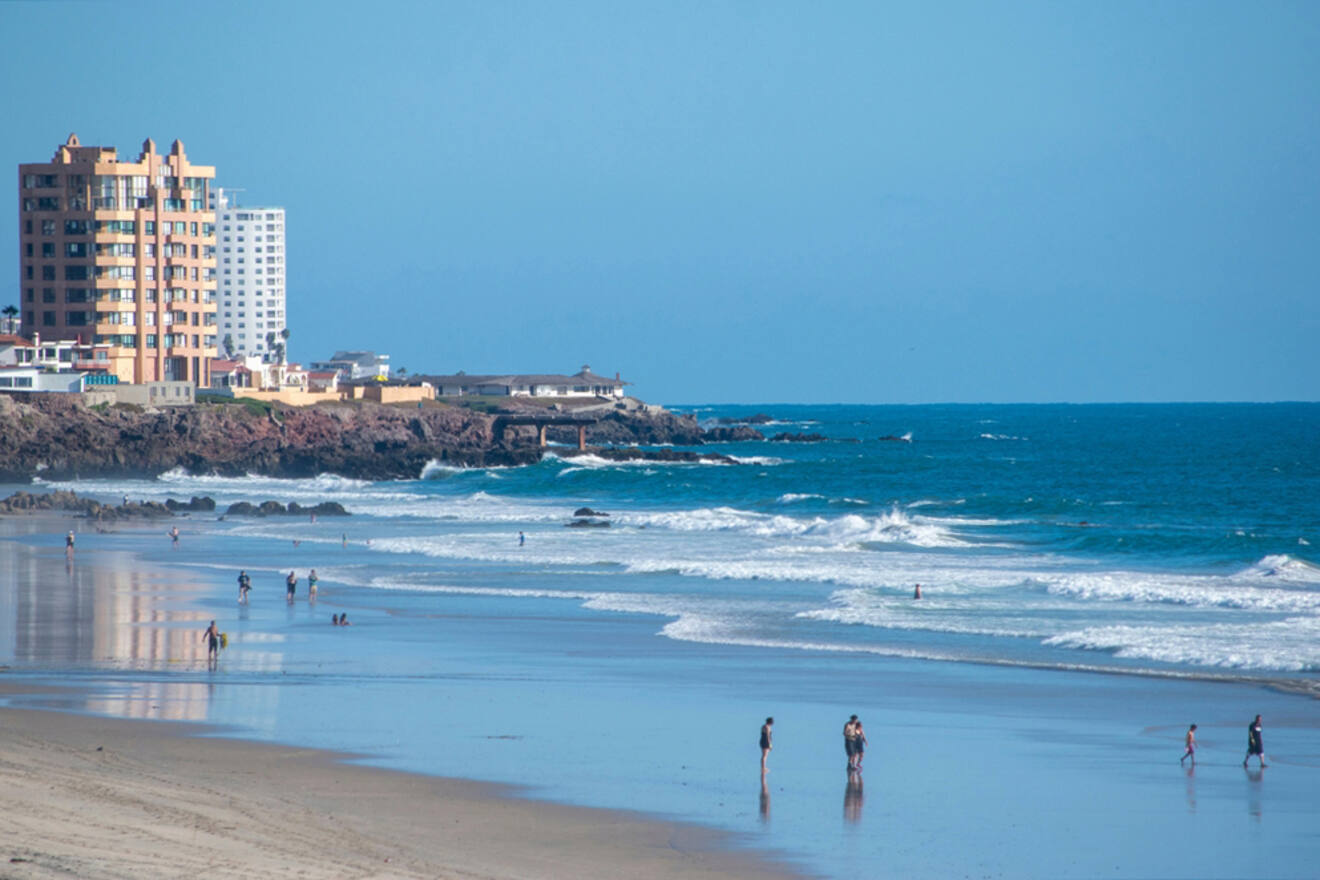 Beach scene with people walking along the shoreline. Waves roll onto the sandy coast. Buildings and rocks are visible in the background under a clear blue sky.