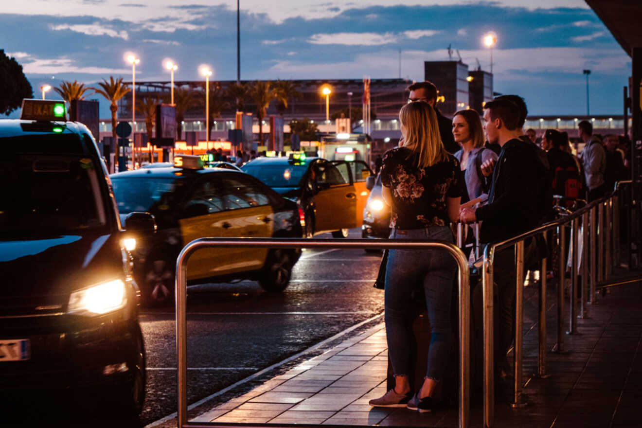 People wait by a railing at a taxi stand during the evening, with several cars and lit streetlights in the background.