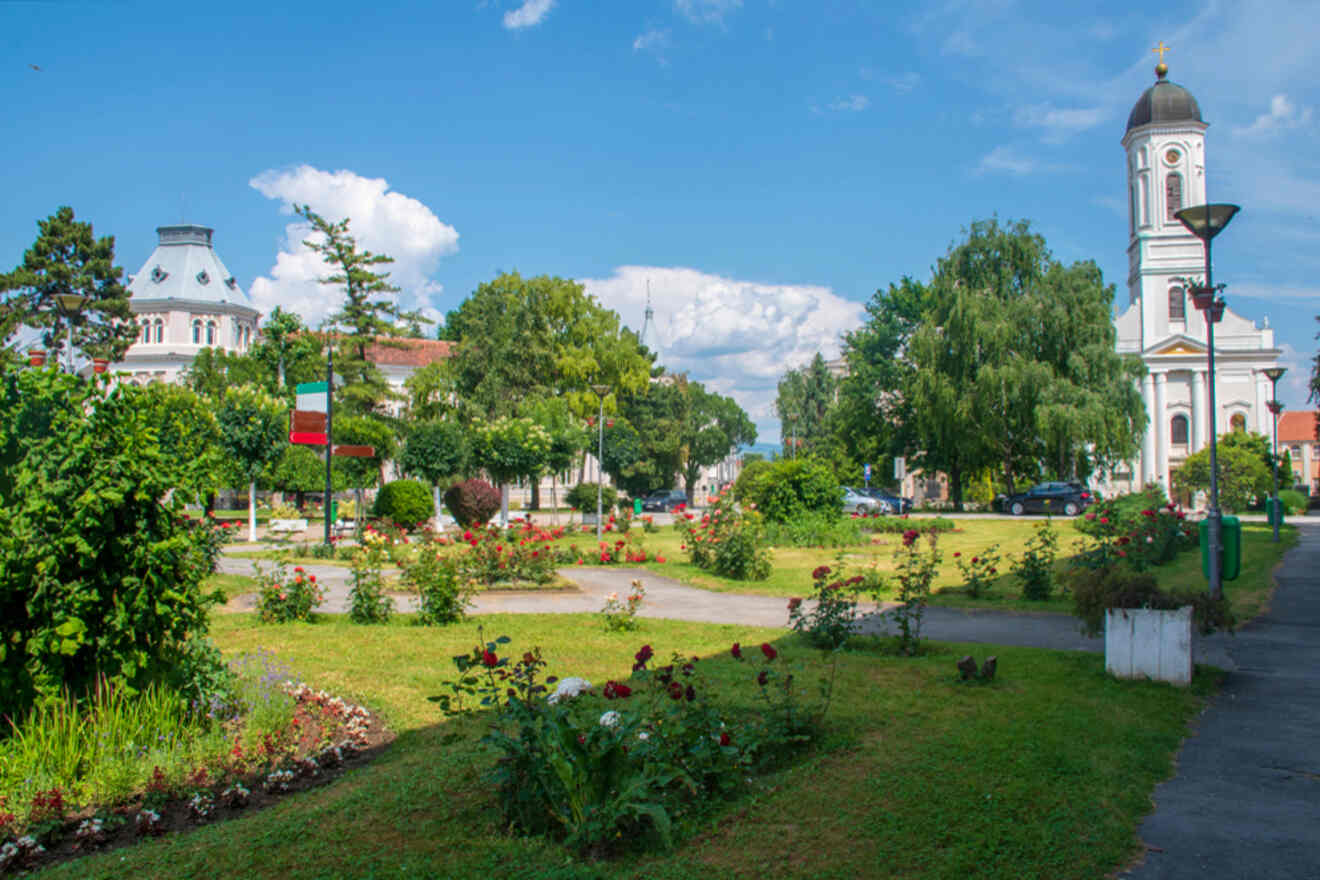 A lush city park with blooming flower beds, a pathway, and a historic church tower in the background under a clear blue sky.