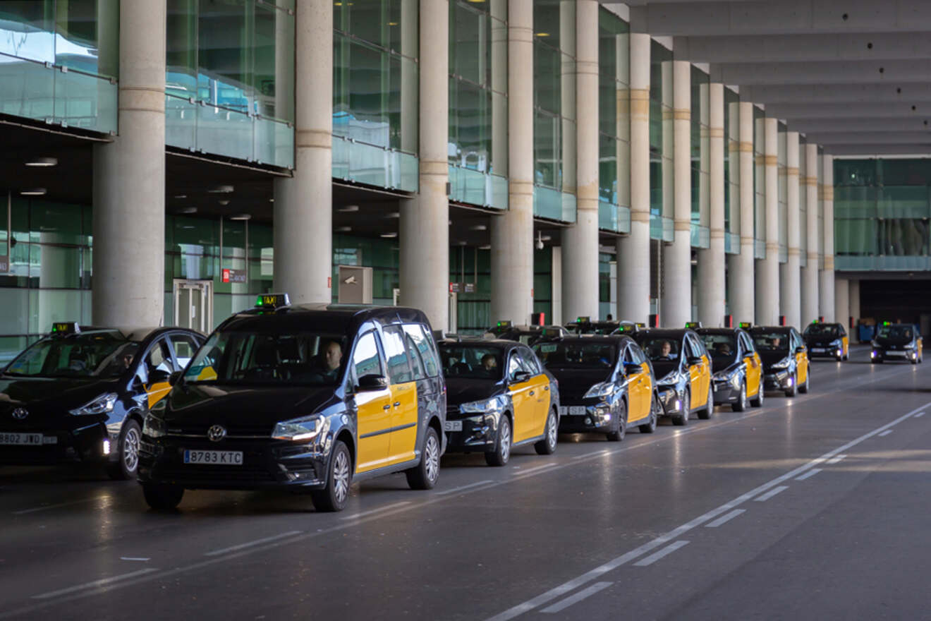 A line of black and yellow taxis wait in a queue under a covered area at an airport terminal.