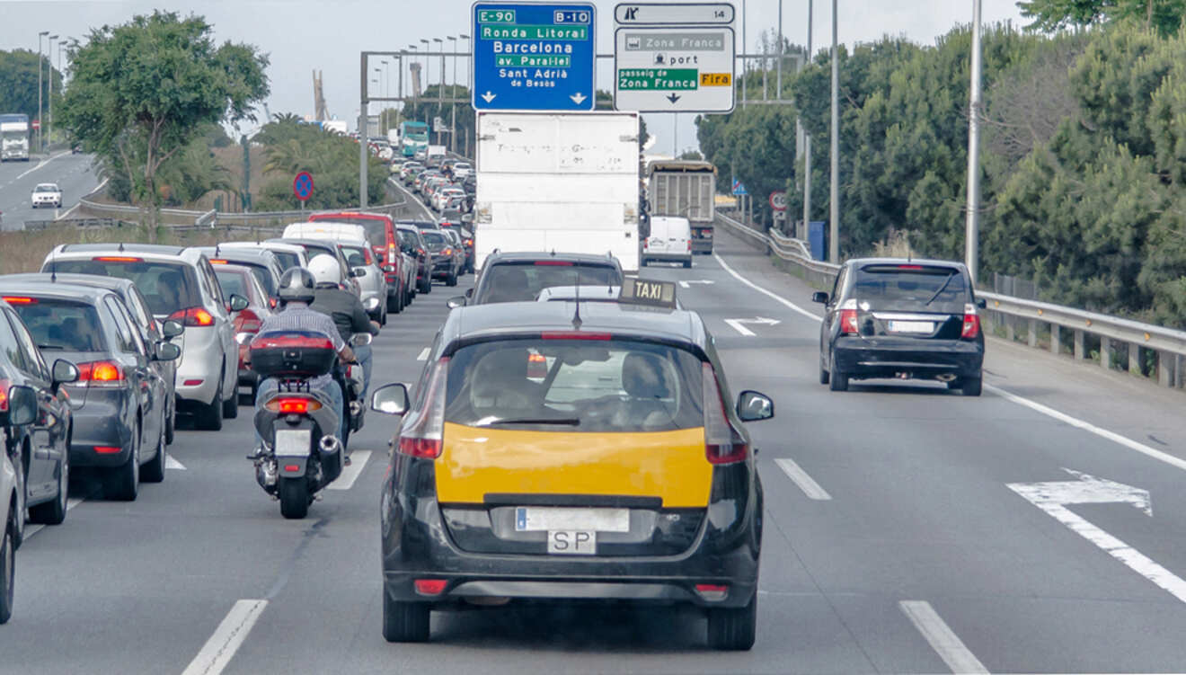 A taxi drives in traffic on a highway near signs for Barcelona. Cars and a motorcycle are on the road.