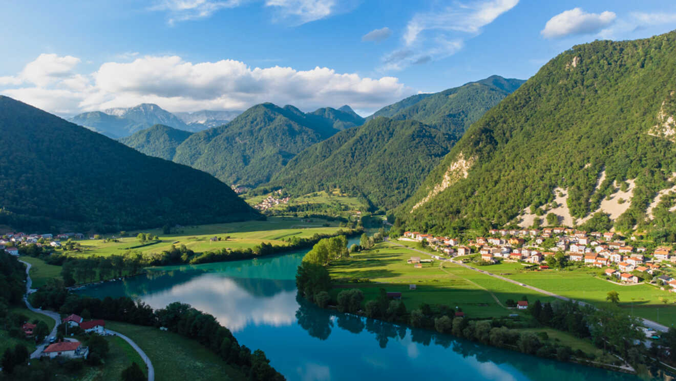 A river flows through a green valley surrounded by forested mountains, with a village on one side and cloudy blue skies above.